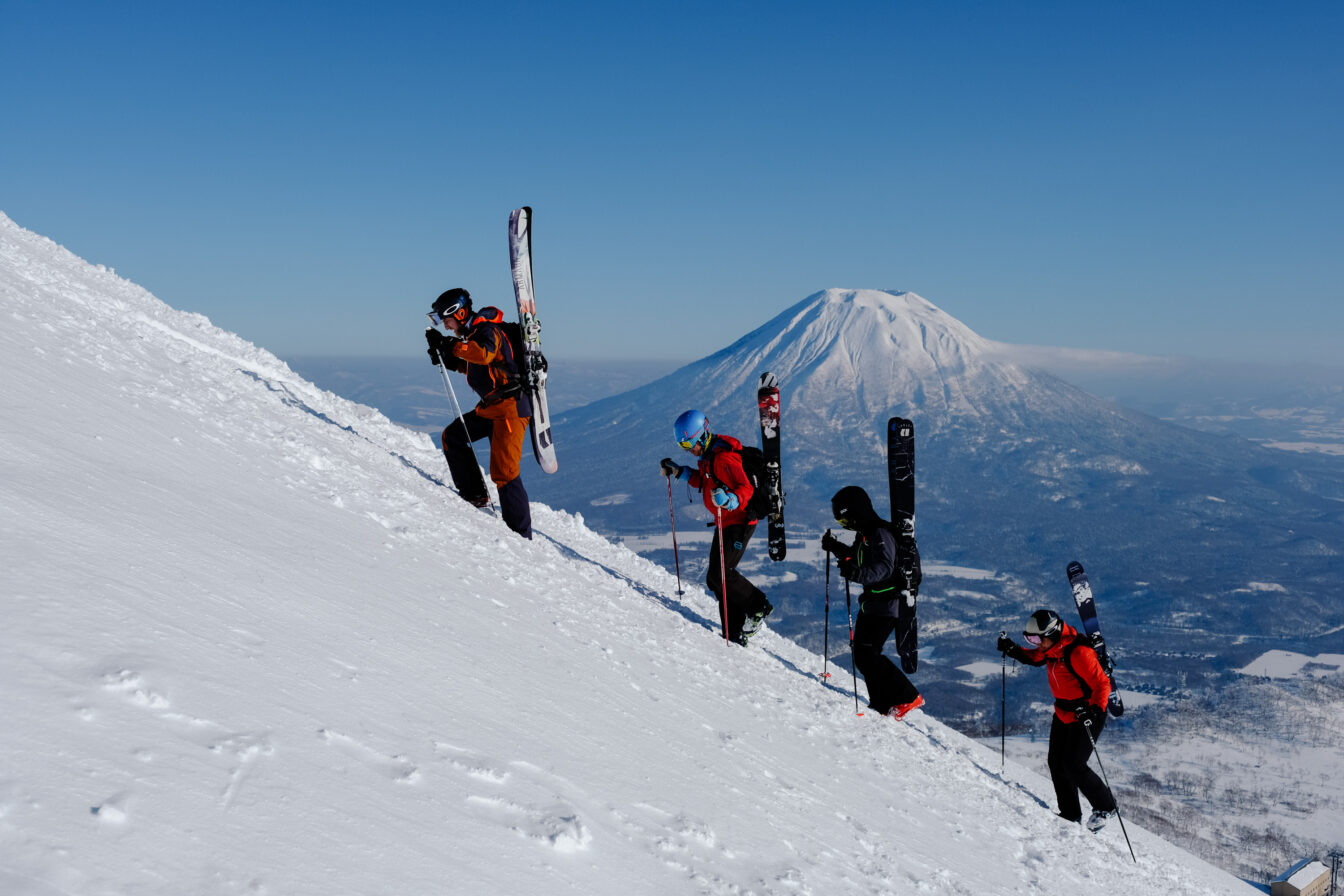 Vier Menschen tragen ihre Ski einen Berg hoch, dahinter ist der schneebedeckte Mount Fuji zu sehen