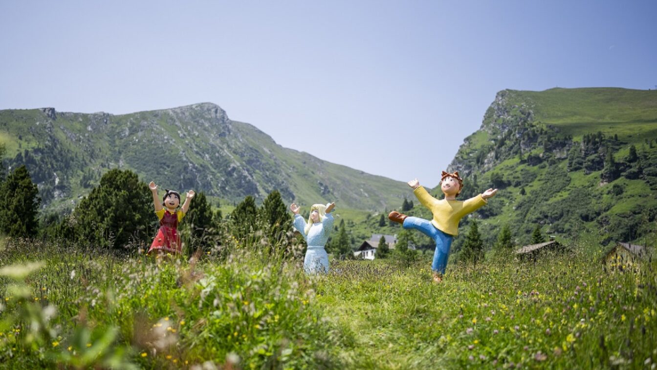 Drei bunte Figuren aus einem Kinderfilm auf einer Blumenwiese mit bergen im Hintergrund