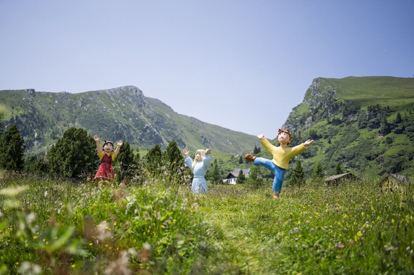 Drei bunte Figuren aus einem Kinderfilm auf einer Blumenwiese mit bergen im Hintergrund