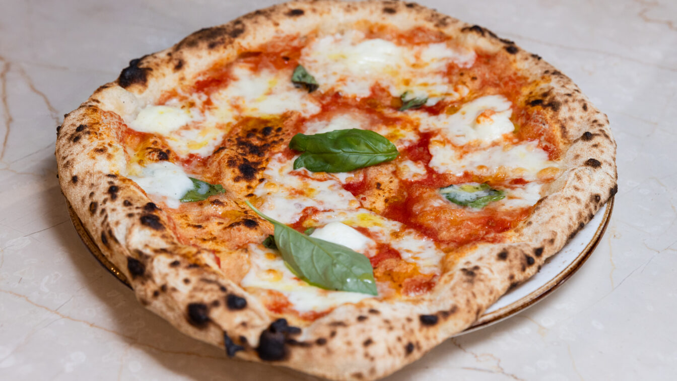 Nahaufnahme einer frisch gebackenen Pizza Margherita mit geschmolzenem Mozzarella, Tomatensauce und frischen Basilikumblättern auf einem hellen Marmortisch.