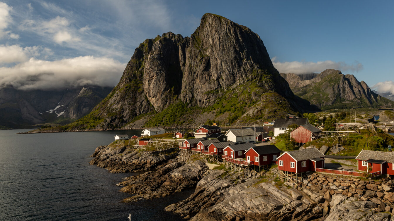 Traditionelle rote Fischerhütten (Rorbuer) in malerischer Küstenlandschaft von Hamnøy auf den Lofoten, Norwegen – mit dramatischer Bergkulisse und klarer Meeresluft unter blauem Himmel.