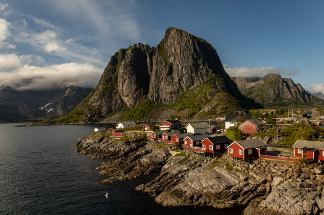 Traditionelle rote Fischerhütten (Rorbuer) in malerischer Küstenlandschaft von Hamnøy auf den Lofoten, Norwegen – mit dramatischer Bergkulisse und klarer Meeresluft unter blauem Himmel.