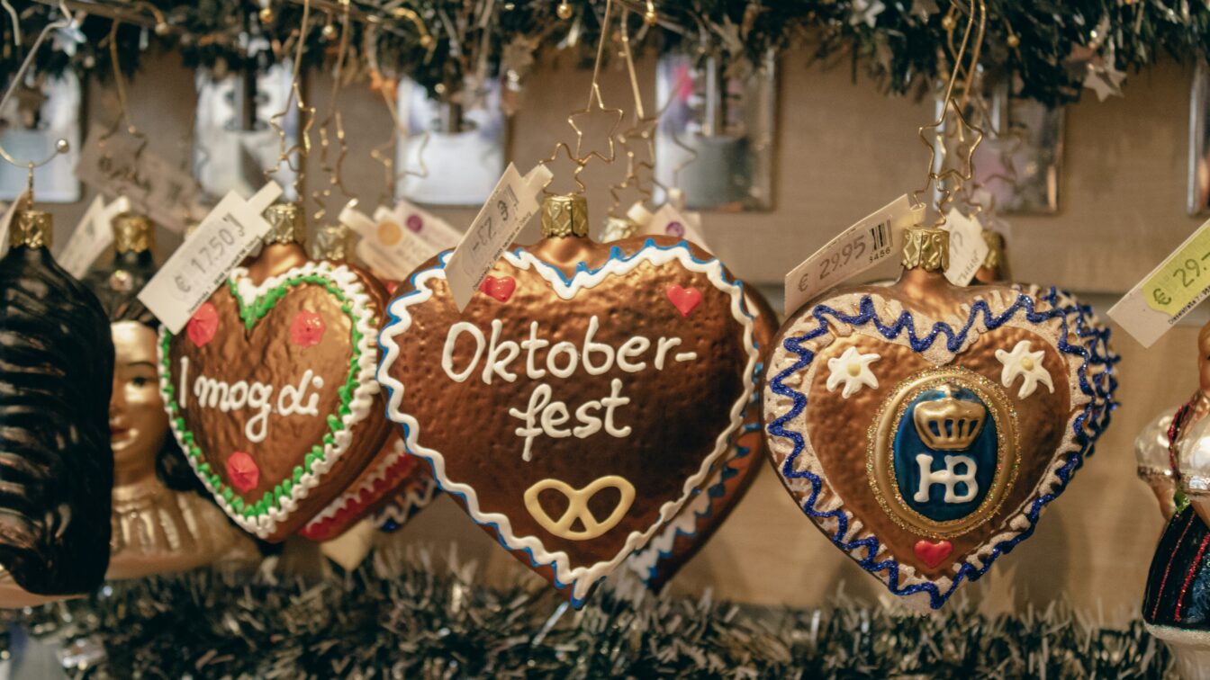 Herzförmige Lebkuchen-Ornamente hängen an Haken in einem festlich dekorierten Verkaufsstand. Die Herzen sind mit buntem Zuckerguss verziert und tragen Aufschriften wie „Oktoberfest“, „I mog di“ und das Hofbräuhaus-Logo „HB“. Preisschilder in Euro sind sichtbar. Im Hintergrund sind Tannengirlanden und weitere weihnachtliche Dekorationen zu sehen.
