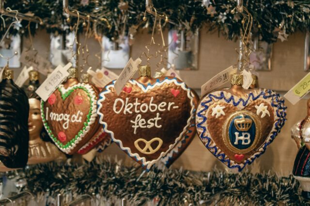 Herzförmige Lebkuchen-Ornamente hängen an Haken in einem festlich dekorierten Verkaufsstand. Die Herzen sind mit buntem Zuckerguss verziert und tragen Aufschriften wie „Oktoberfest“, „I mog di“ und das Hofbräuhaus-Logo „HB“. Preisschilder in Euro sind sichtbar. Im Hintergrund sind Tannengirlanden und weitere weihnachtliche Dekorationen zu sehen.