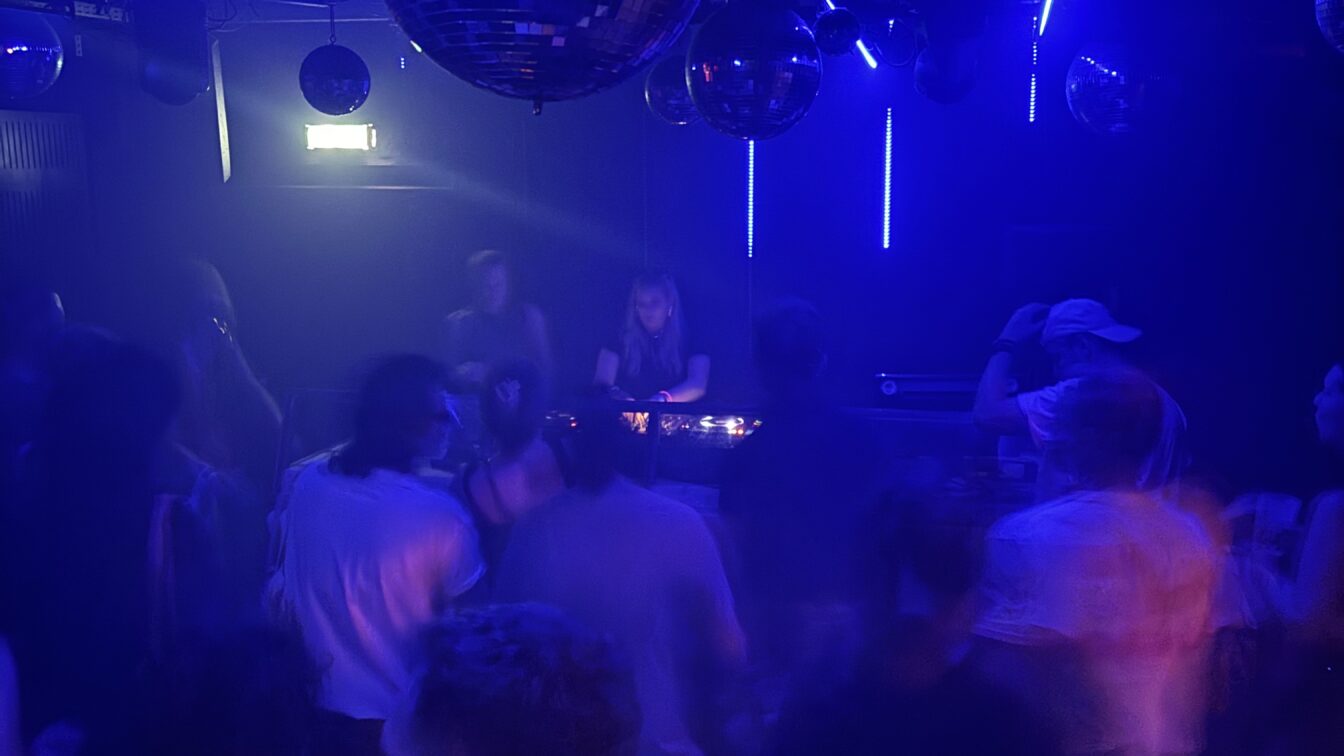 Eine Szene aus einem belebten Nachtclub in Europa: Menschen tanzen unter blauem und violettem Licht, während eine DJane im Hintergrund Musik auflegt. Mehrere Diskokugeln hängen von der Decke und reflektieren das Licht, was eine lebhafte und energiegeladene Atmosphäre schafft. Die Bewegungen der Menschen wirken leicht verschwommen, was auf Tanzen und Bewegung hindeutet.