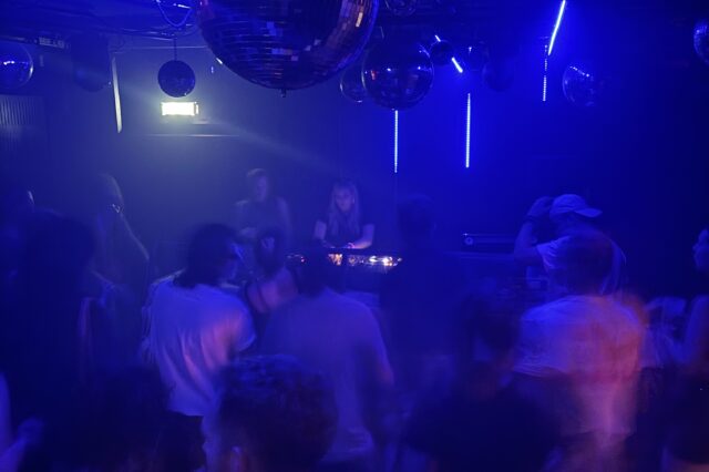 Eine Szene aus einem belebten Nachtclub in Europa: Menschen tanzen unter blauem und violettem Licht, während eine DJane im Hintergrund Musik auflegt. Mehrere Diskokugeln hängen von der Decke und reflektieren das Licht, was eine lebhafte und energiegeladene Atmosphäre schafft. Die Bewegungen der Menschen wirken leicht verschwommen, was auf Tanzen und Bewegung hindeutet.