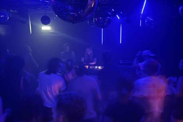 Eine Szene aus einem belebten Nachtclub in Europa: Menschen tanzen unter blauem und violettem Licht, während eine DJane im Hintergrund Musik auflegt. Mehrere Diskokugeln hängen von der Decke und reflektieren das Licht, was eine lebhafte und energiegeladene Atmosphäre schafft. Die Bewegungen der Menschen wirken leicht verschwommen, was auf Tanzen und Bewegung hindeutet.