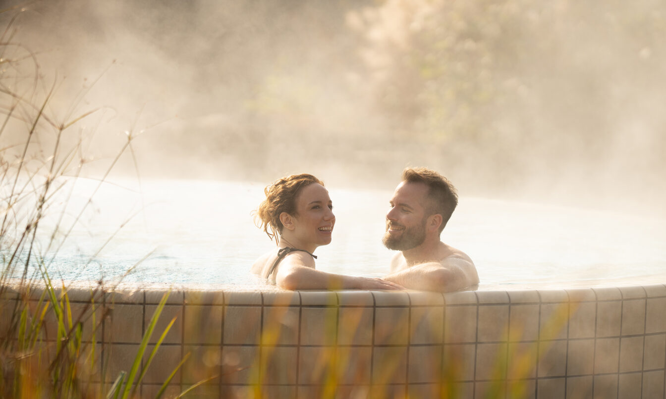 Eine Frau und ein Mann entspannen bei Abendsonne in einem dampfenden Thermalpool.
