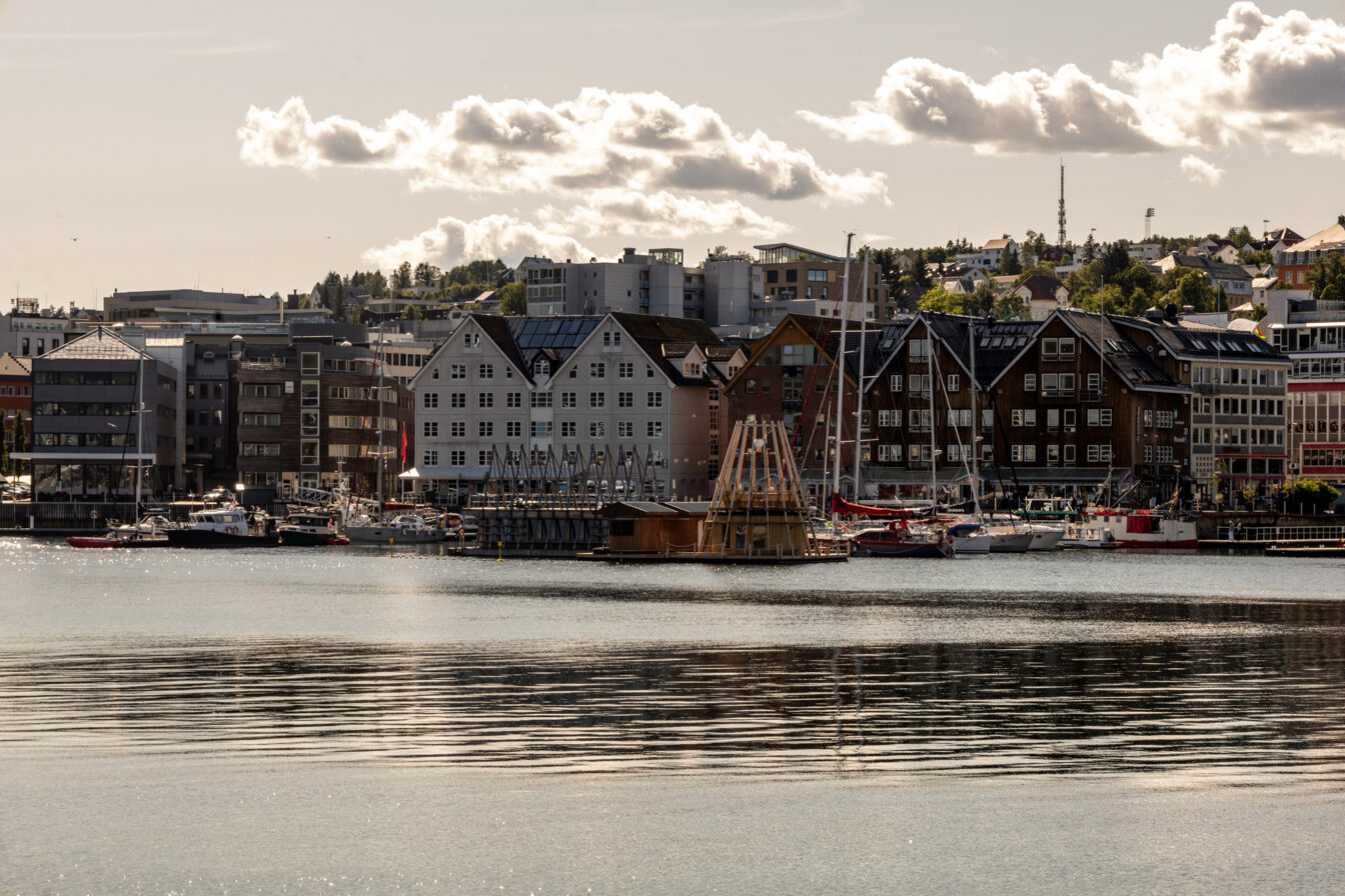 Blick über das Wasser auf eine schwimmende Sauna mit spitz zulaufender Holzarchitektur im Hafen von Tromsø. Dahinter liegen moderne und traditionelle Hafengebäude mit Giebeldächern sowie zahlreiche Segelboote. Die Szene ist in warmes Sonnenlicht getaucht, darüber ein Himmel mit dramatisch geformten Wolken.