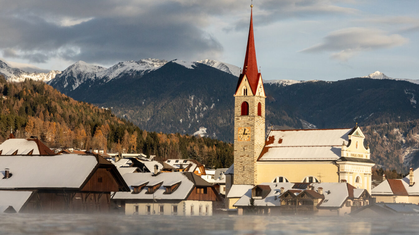 Winterliches Südtiroler Dorf mit schneebedeckten Dächern, einer Kirche mit rotem Turm und umliegenden Alpenbergen im Hintergrund bei warmem Abendlicht. Im Vordergrund sieht man das Sky Pool vom Hotel Solvie im Pustertal