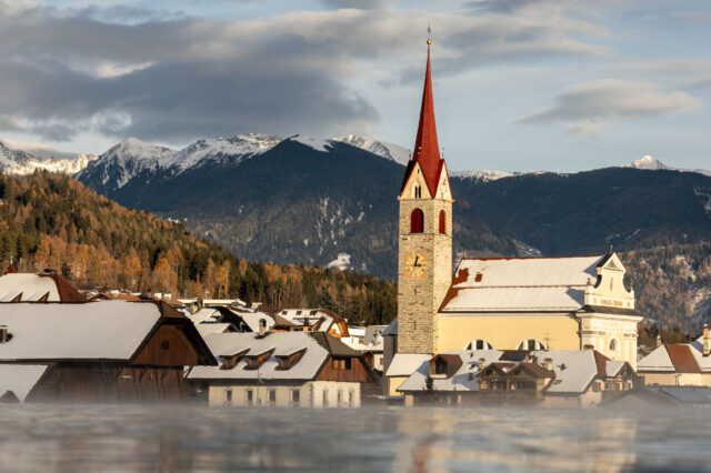 Winterliches Südtiroler Dorf mit schneebedeckten Dächern, einer Kirche mit rotem Turm und umliegenden Alpenbergen im Hintergrund bei warmem Abendlicht. Im Vordergrund sieht man das Sky Pool vom Hotel Solvie im Pustertal