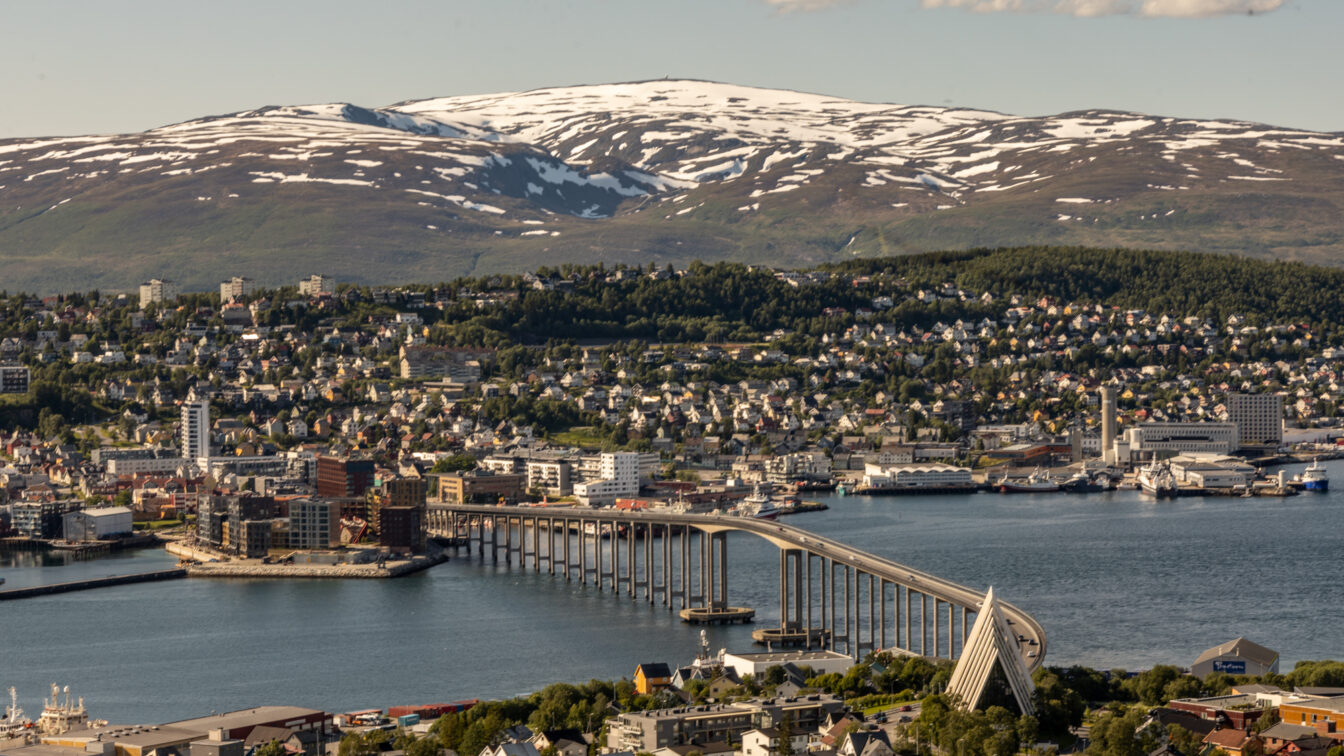 Luftaufnahme der Stadt Tromsø in Norwegen mit dem markanten Tromsø-Brücke, die sich über den Fjord erstreckt. Im Vordergrund ist die moderne Eismeerkathedrale zu sehen, im Hintergrund schneebedeckte Berge unter blauem Himmel mit vereinzelten Wolken.