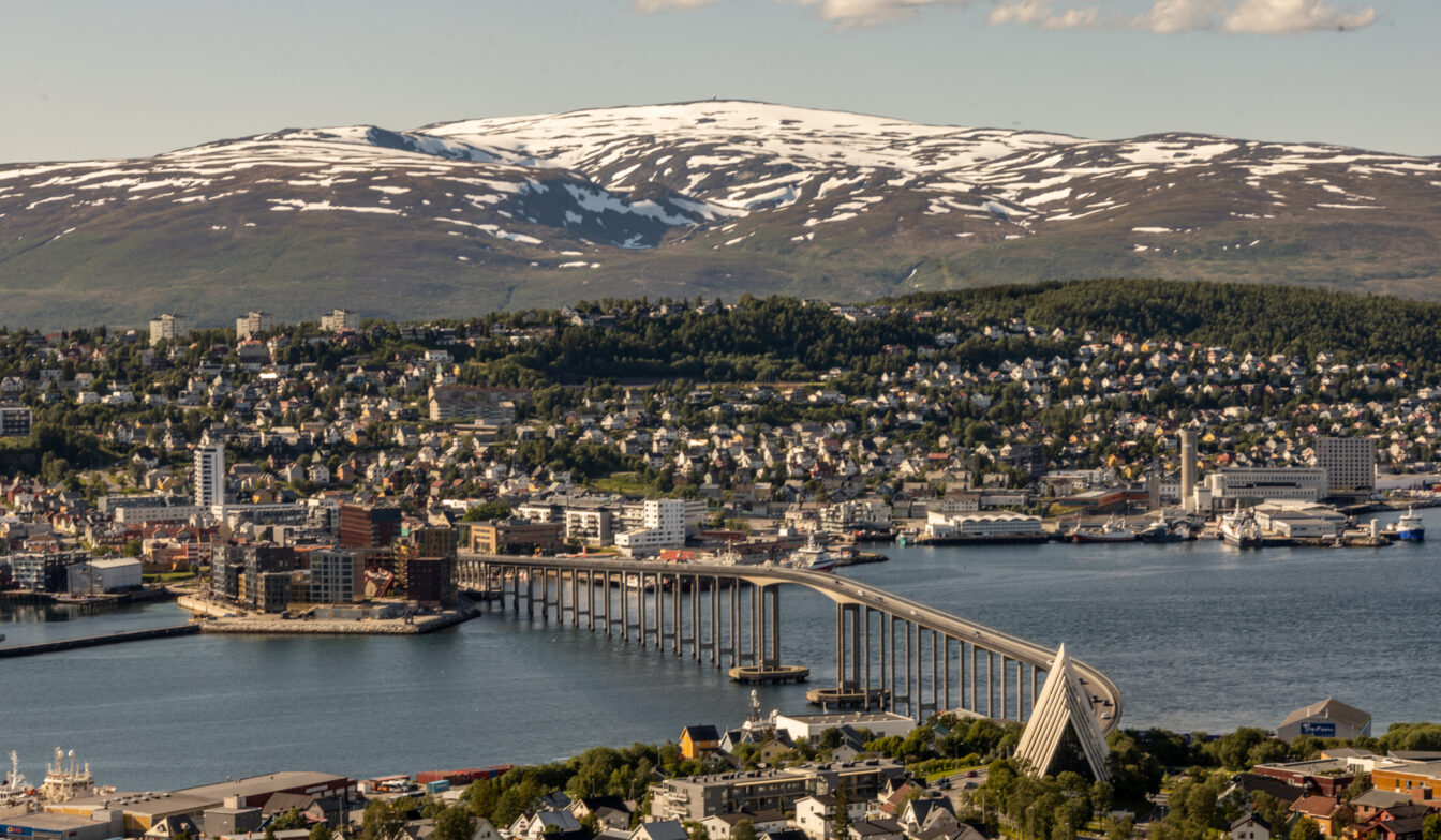 Luftaufnahme der Stadt Tromsø in Norwegen mit dem markanten Tromsø-Brücke, die sich über den Fjord erstreckt. Im Vordergrund ist die moderne Eismeerkathedrale zu sehen, im Hintergrund schneebedeckte Berge unter blauem Himmel mit vereinzelten Wolken.
