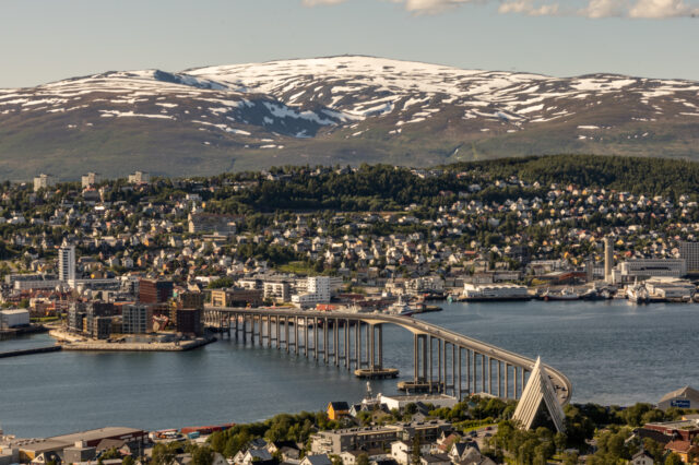 Luftaufnahme der Stadt Tromsø in Norwegen mit dem markanten Tromsø-Brücke, die sich über den Fjord erstreckt. Im Vordergrund ist die moderne Eismeerkathedrale zu sehen, im Hintergrund schneebedeckte Berge unter blauem Himmel mit vereinzelten Wolken.