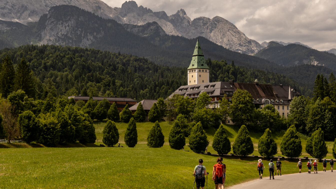 Wanderer auf einem Weg zum Schloss Elmau bei Garmisch-Partenkirchen, umgeben von grüner Natur und den majestätischen Alpen.