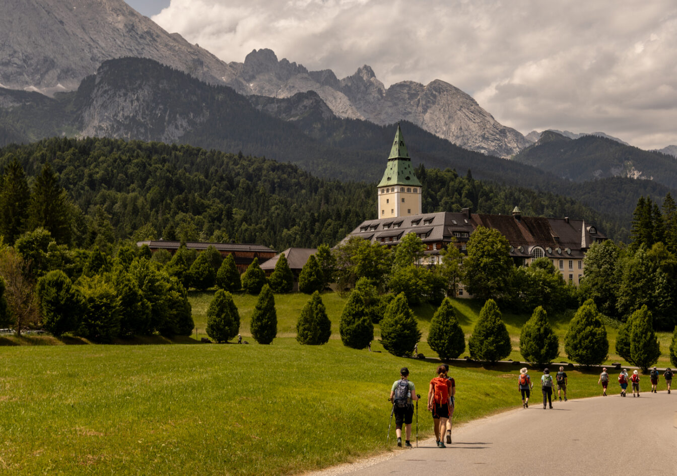Wanderer auf einem Weg zum Schloss Elmau bei Garmisch-Partenkirchen, umgeben von grüner Natur und den majestätischen Alpen.