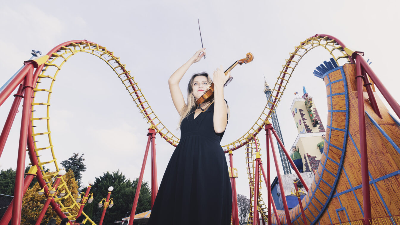 Eine Frau in einem langen schwarzen Kleid spielt Geige im Wiener Prater vor einer markanten rot-gelben Looping-Achterbahn. Im Hintergrund sind weitere Fahrgeschäfte und ein Turm zu sehen. Um sie herum stehen Besucher. Die Szene stammt vom ISA Festival in Wien, das kostenlose Konzerte in ganz Wien bietet.