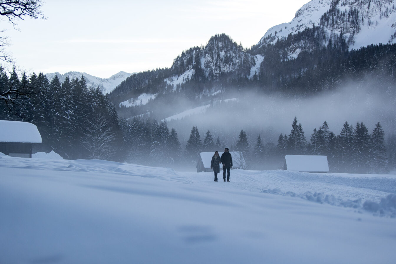 Zwei Personen wandern durch den Schnee, im Hintergrund ist ein Bergpanorama zu sehen.