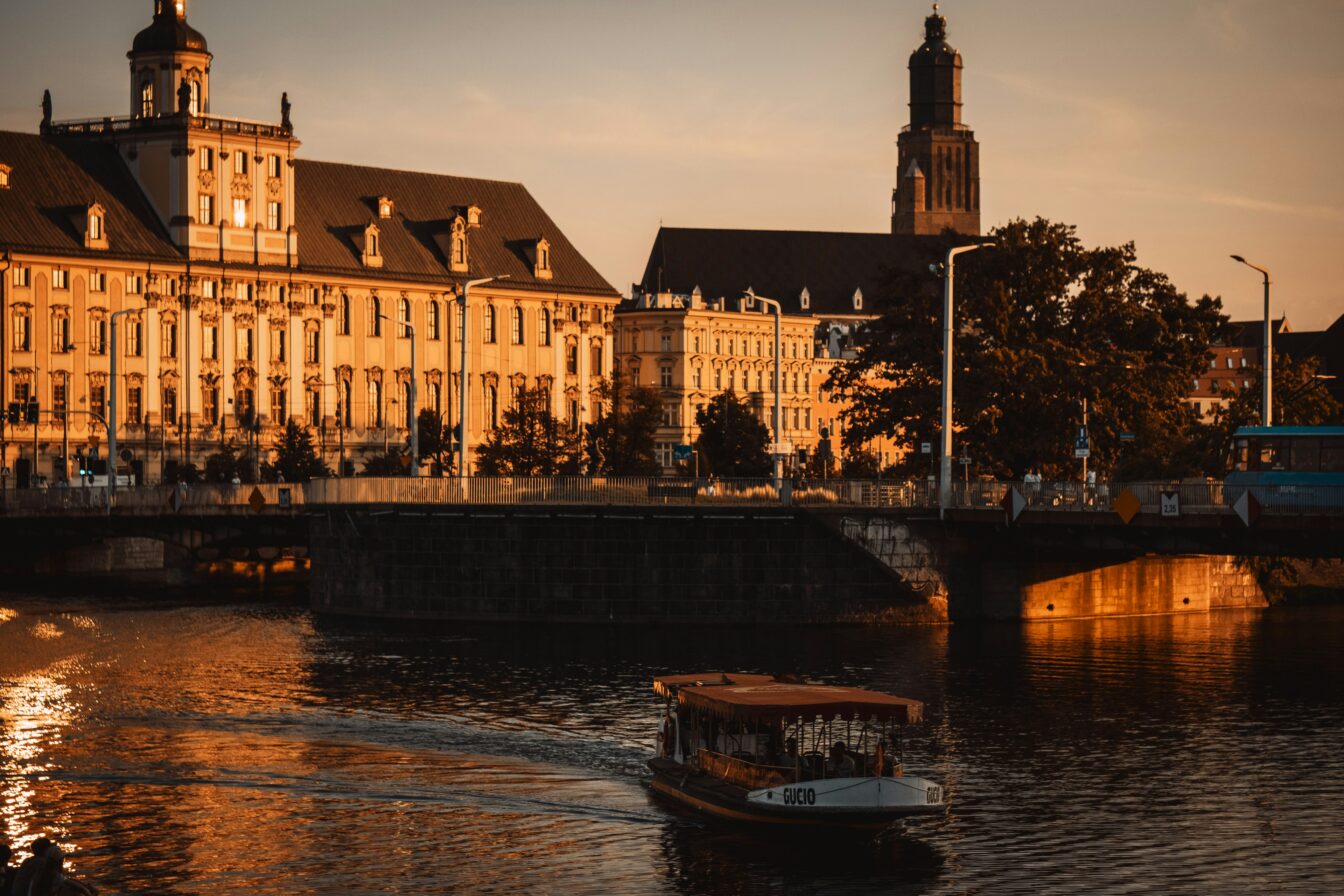Ein Boot fährt bei goldenem Sonnenuntergang auf der Oder in Wrocław entlang, während historische Gebäude und eine Brücke im warmen Licht leuchten.