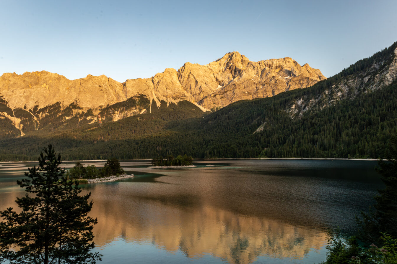 Sonnenuntergang am Eibsee mit Blick auf die Zugspitze in Garmisch-Partenkirchen, der höchste Berg Deutschlands leuchtet in warmem Licht, während sich die Bergspitzen im ruhigen, türkisfarbenen Wasser des Sees spiegeln, umgeben von dichten Wäldern.