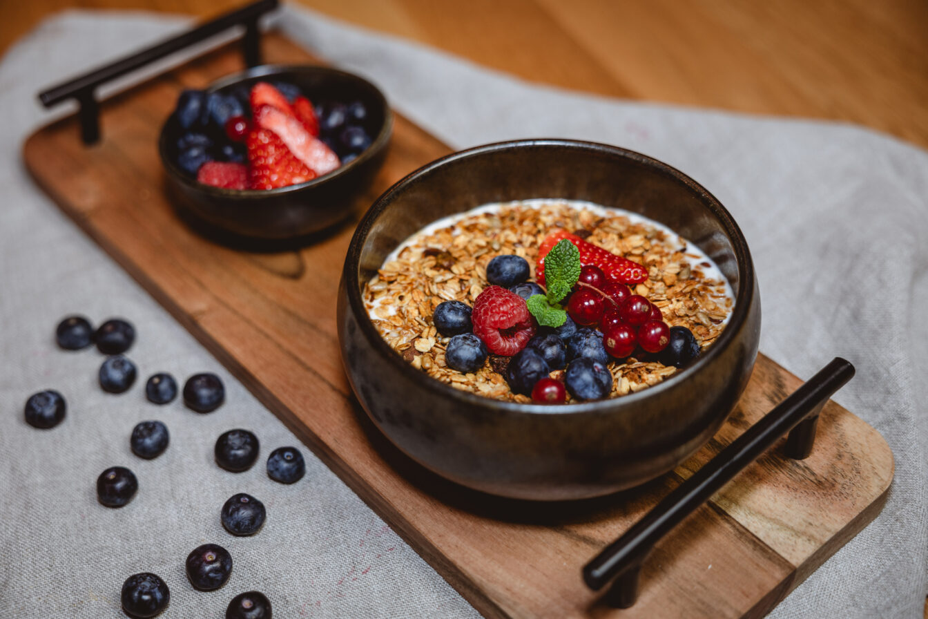Eine Schale mit Joghurt, Müsli und frischen Beeren auf einem Tablett aus Holz.