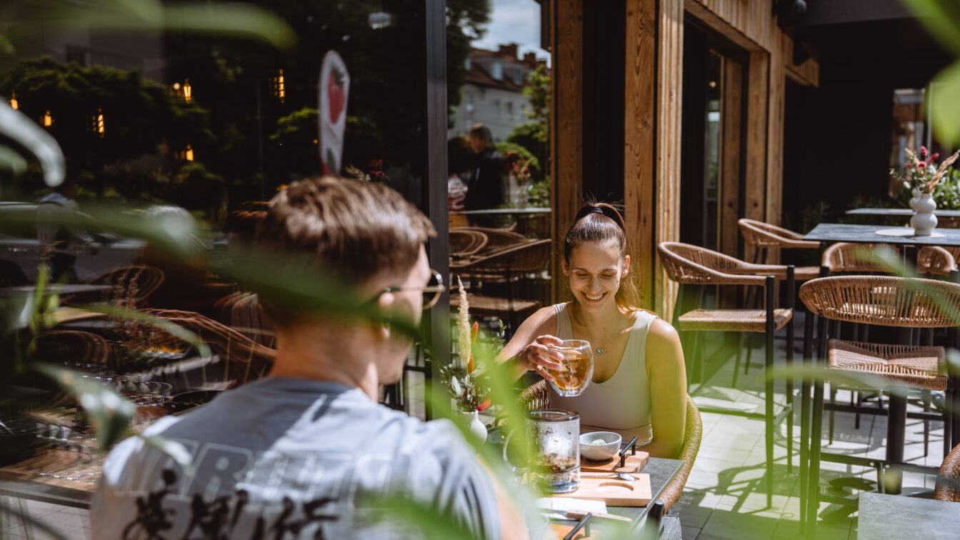 Zwei Menschen auf einer Terrasse, die begrünt und sonnig ist.