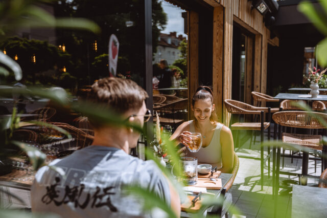 Zwei Menschen auf einer Terrasse, die begrünt und sonnig ist.
