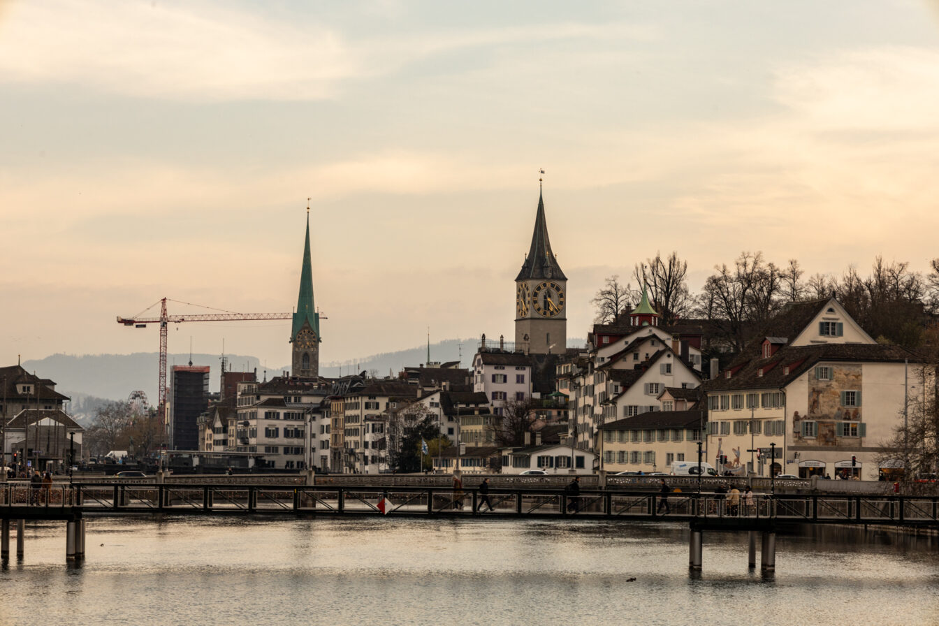 Blick auf das Panorama einer Stadt mit Fluss im Vordergrund und Bergen im Hintergrund.