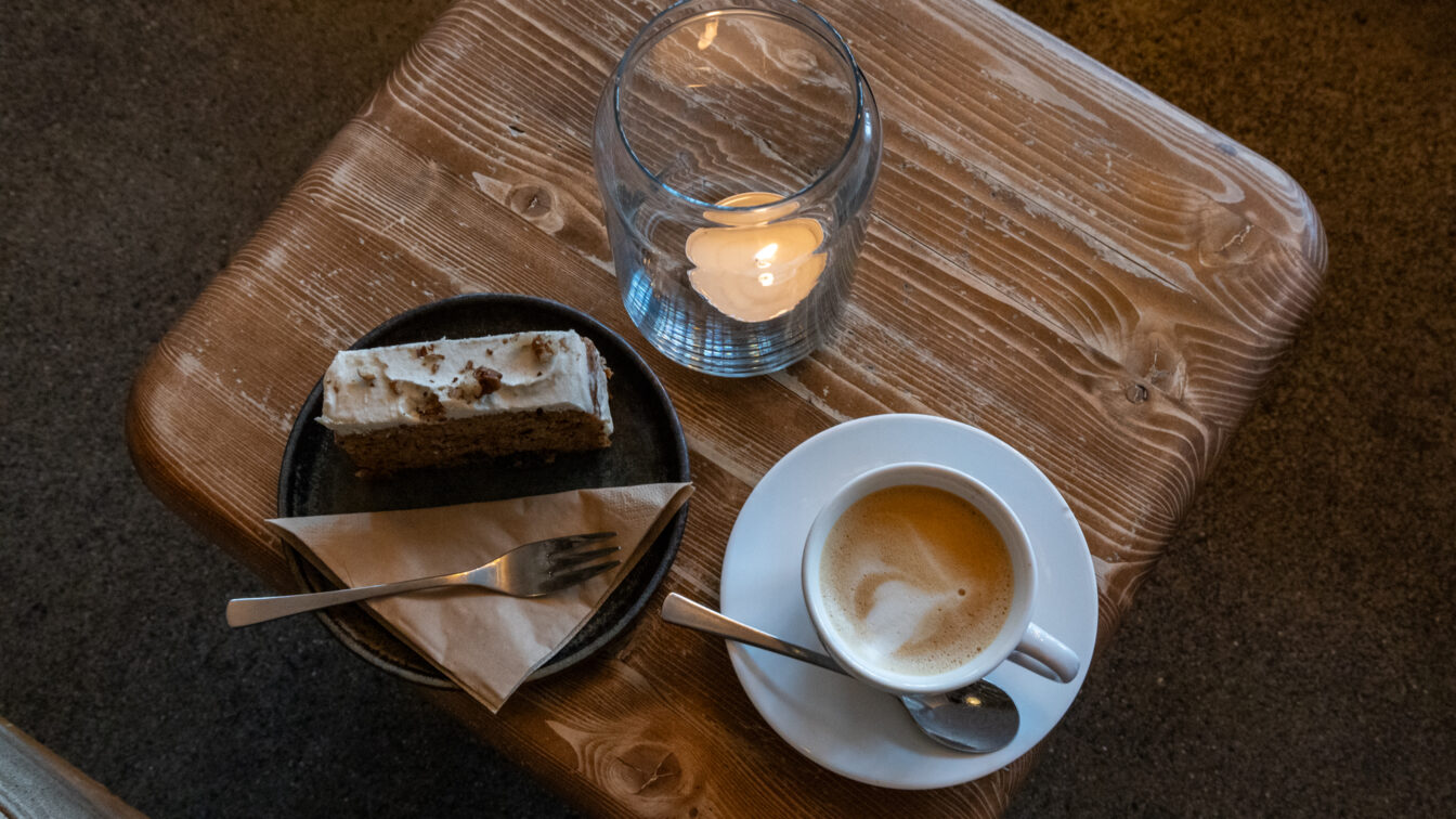Eine Tasse Kaffee mit Milchschaum, ein Stück Kuchen auf einem Teller und eine brennende Kerze auf einem rustikalen Holztisch in gemütlicher Café-Atmosphäre in Berlin