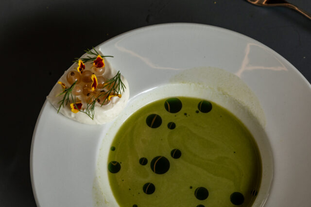 Fein angerichtetes Gericht in einem Restaurant in Berlin-Mitte: Eine grüne Suppe mit dunklen Ölkreisen, serviert in einem weißen Teller. Am Rand eine cremige Garnitur mit hellen Beeren, frischem Dill und essbaren Blüten.
