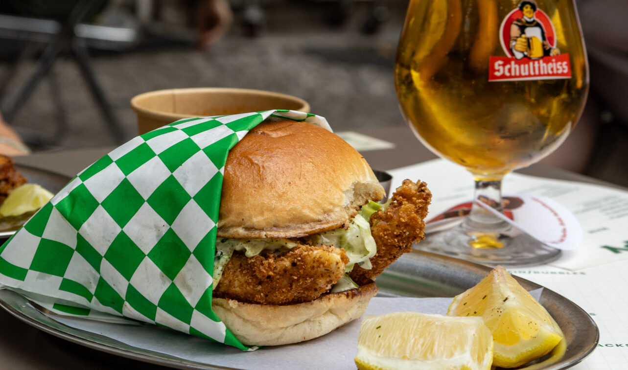 Knuspriger Fischburger mit Zitronenspalten und frisch gezapftem Schultheiss-Bier auf einem Tisch in Berlin, aufgenommen bei einer Sommer-Neueröffnung 2025, im Hintergrund entspannte Gäste in geselliger Atmosphäre.
