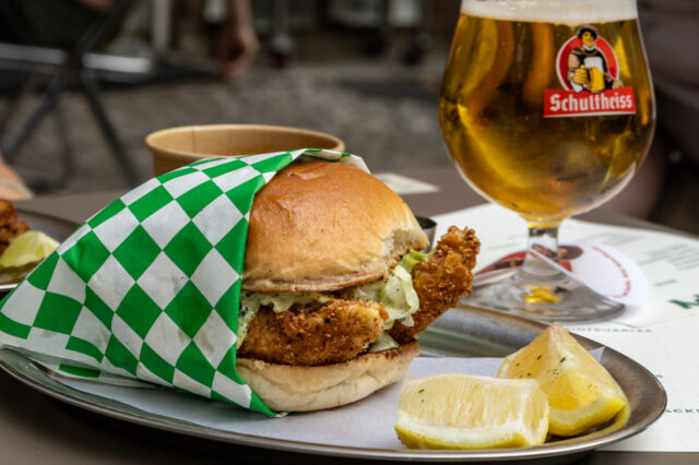 Knuspriger Fischburger mit Zitronenspalten und frisch gezapftem Schultheiss-Bier auf einem Tisch in Berlin, aufgenommen bei einer Sommer-Neueröffnung 2025, im Hintergrund entspannte Gäste in geselliger Atmosphäre.