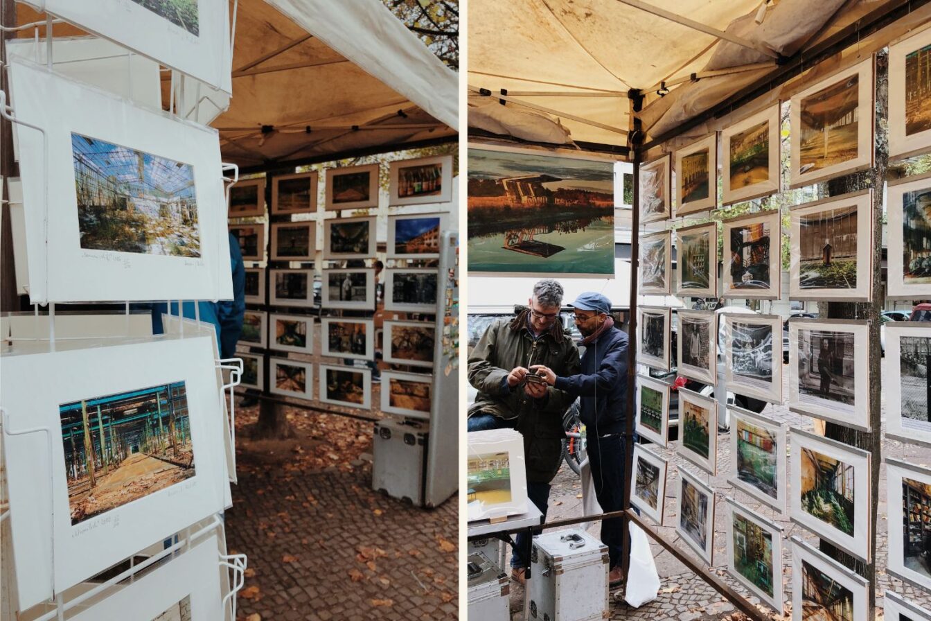Fotostände mit Kunstdrucken auf dem Flohmarkt Boxhagener Platz in Berlin-Friedrichshain – zwei Männer betrachten gemeinsam ein Smartphone zwischen den ausgestellten Bildern unter einem Marktstand