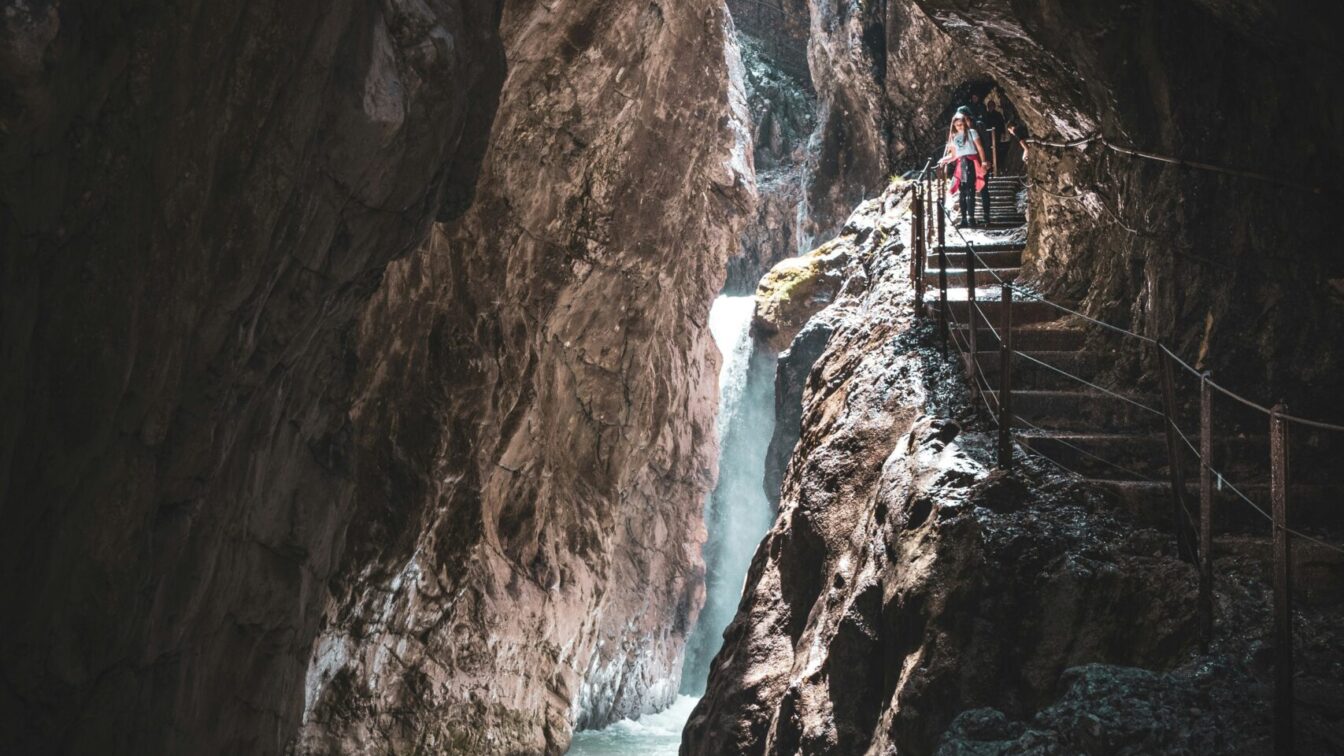 Schmale Felsenschlucht der Höllentalklamm mit rauschendem Wildbach und Wasserfall, ein schmaler Steg mit Geländer führt an der Felswand entlang, zwei Wanderer gehen darauf, während Lichtstrahlen zwischen den steilen Felswänden hindurchfallen.