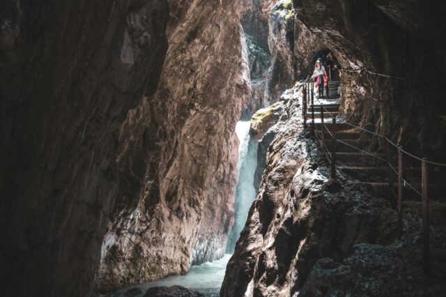Schmale Felsenschlucht der Höllentalklamm mit rauschendem Wildbach und Wasserfall, ein schmaler Steg mit Geländer führt an der Felswand entlang, zwei Wanderer gehen darauf, während Lichtstrahlen zwischen den steilen Felswänden hindurchfallen.
