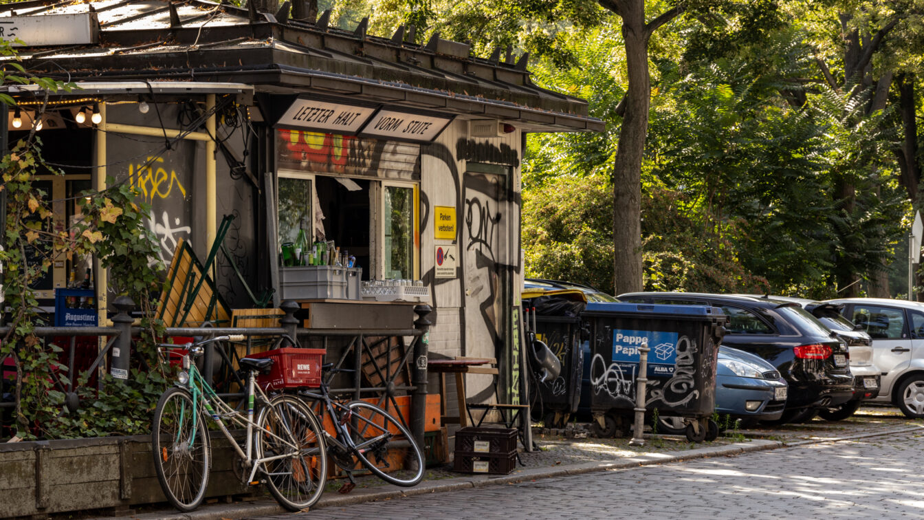 Straßenszene in Berlin-Kreuzberg mit einem kleinen, mit Graffiti verzierten Kiosk, Fahrrädern, Müllcontainern und parkenden Autos unter grünen Bäumen bei Sonnenschein.