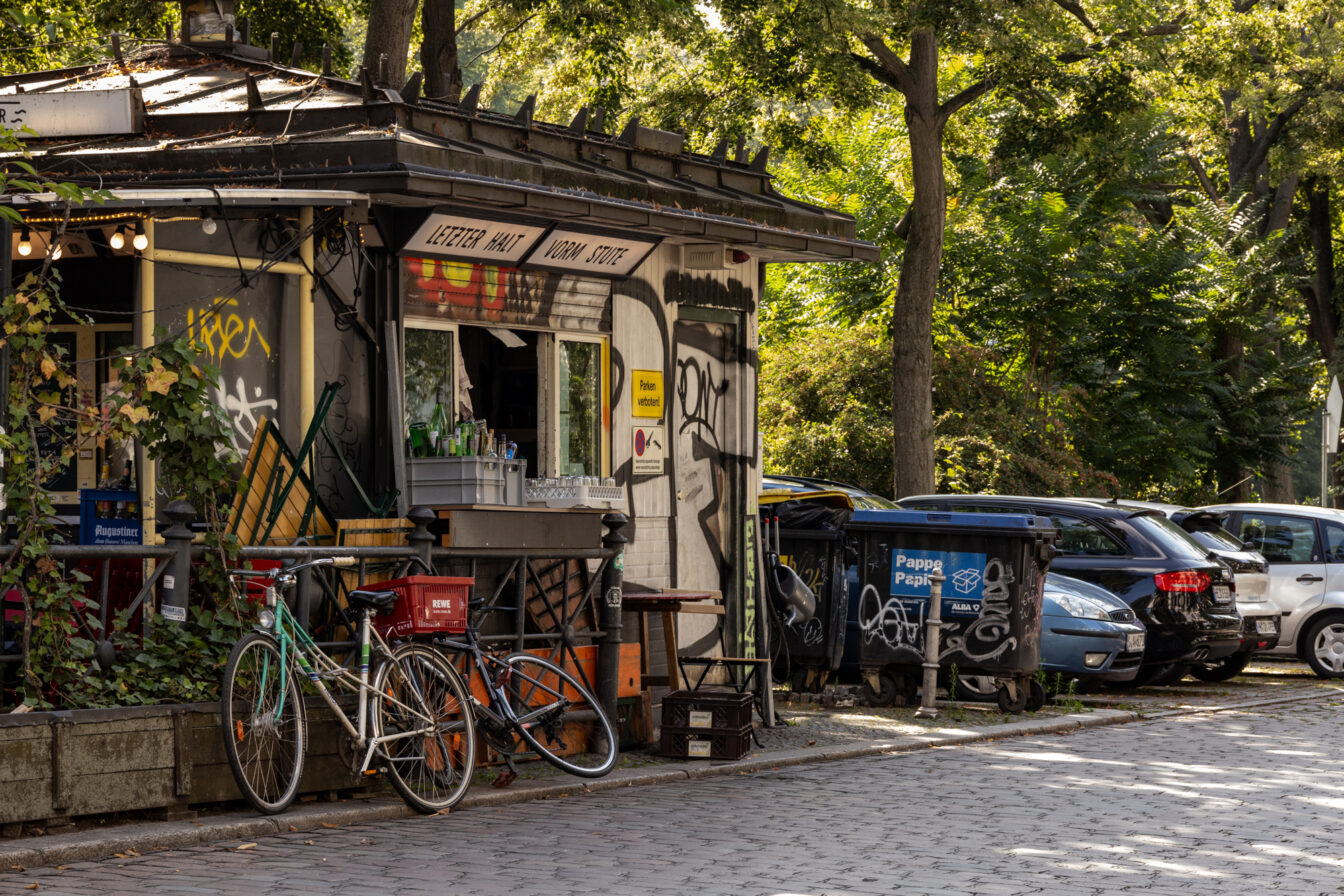 Straßenszene in Berlin-Kreuzberg mit einem kleinen, mit Graffiti verzierten Kiosk, Fahrrädern, Müllcontainern und parkenden Autos unter grünen Bäumen bei Sonnenschein.