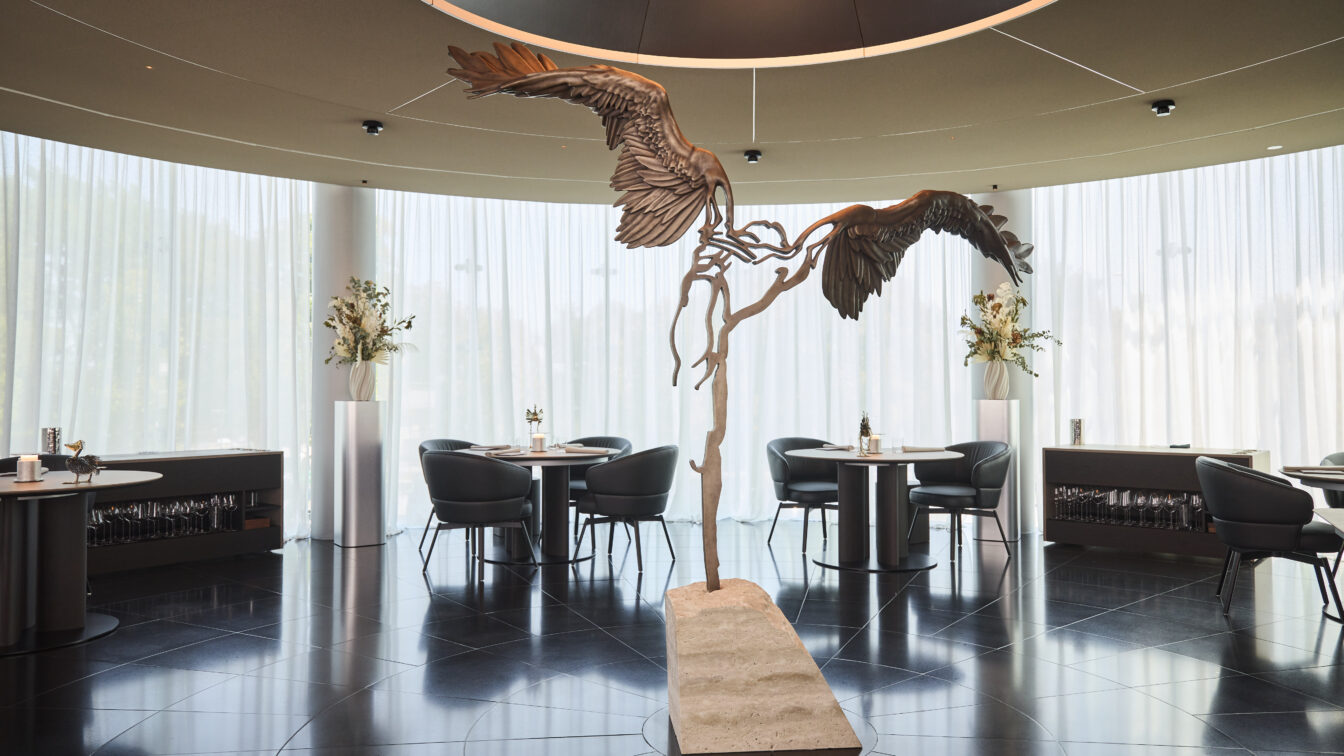Elegantes Fine-Dining-Restaurant mit moderner, runder Architektur und hohen, lichtdurchlässigen Vorhängen. Im Zentrum steht eine kunstvolle Skulptur mit ausgebreiteten Flügeln, die an den Ikarus-Mythos erinnert. Die Einrichtung ist stilvoll mit runden Tischen, dunklen Sesseln und dezenten Blumendekorationen.