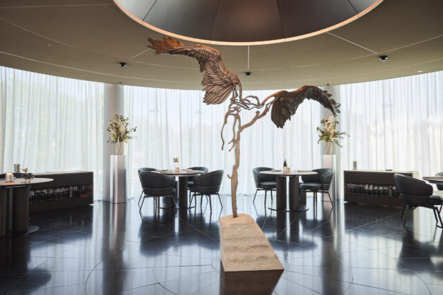 Elegantes Fine-Dining-Restaurant mit moderner, runder Architektur und hohen, lichtdurchlässigen Vorhängen. Im Zentrum steht eine kunstvolle Skulptur mit ausgebreiteten Flügeln, die an den Ikarus-Mythos erinnert. Die Einrichtung ist stilvoll mit runden Tischen, dunklen Sesseln und dezenten Blumendekorationen.