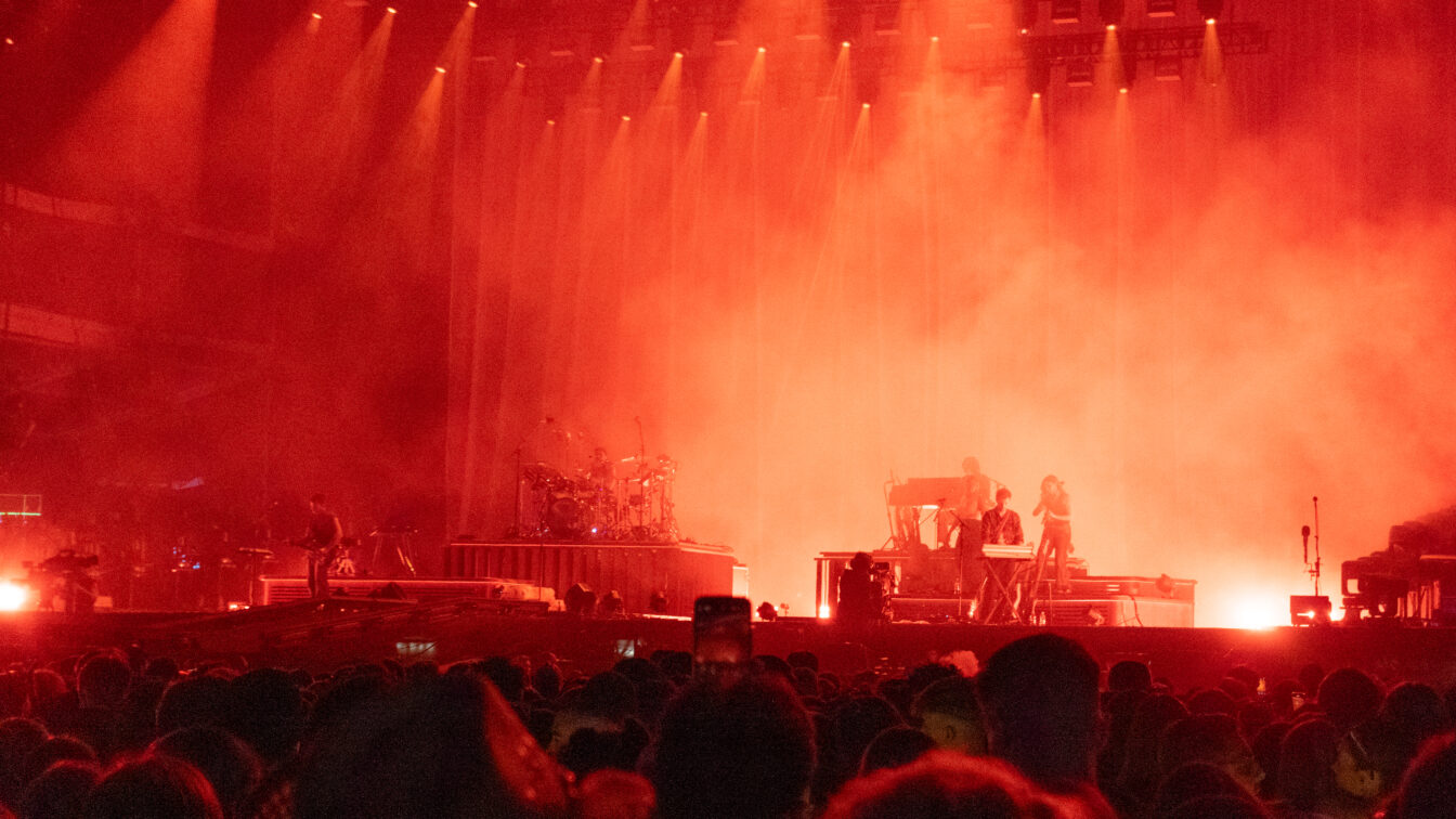 Bühne bei einem Open-Air-Konzert in Berlin, in rotes Licht getaucht. Musikerinnen und Musiker spielen auf Instrumenten wie Schlagzeug, Keyboard und Gitarre, während dichtes Publikum davor steht. Nebel und Scheinwerfer verstärken die intensive Atmosphäre.