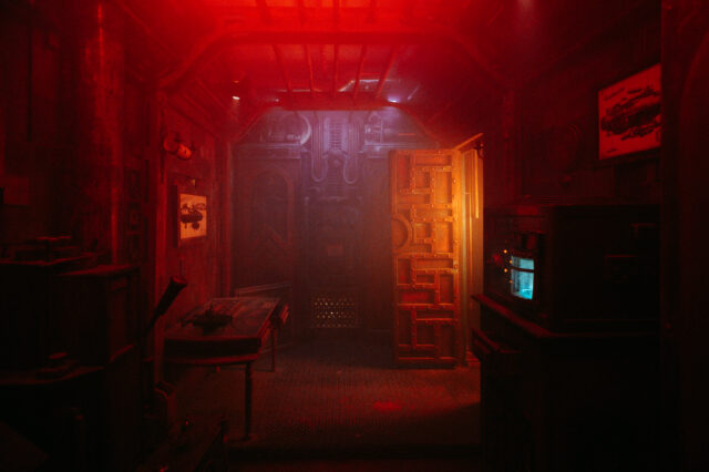 Ein schmaler, düster beleuchteter Raum mit industriellem und futuristischem Design, in rotem Licht getaucht. An den Wänden hängen technische Zeichnungen und Metallrohre verlaufen an der Decke. Rechts befindet sich ein Gerät mit leuchtendem blauen Bildschirm. Eine massive Tür aus Metall und Holz ist halb geöffnet und führt in einen weiteren, noch dunkleren Bereich.