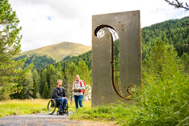 Eine Person im Rollstuhl und eine nicht beeinträchtigte Person vor einer Skulptur im Grünen.