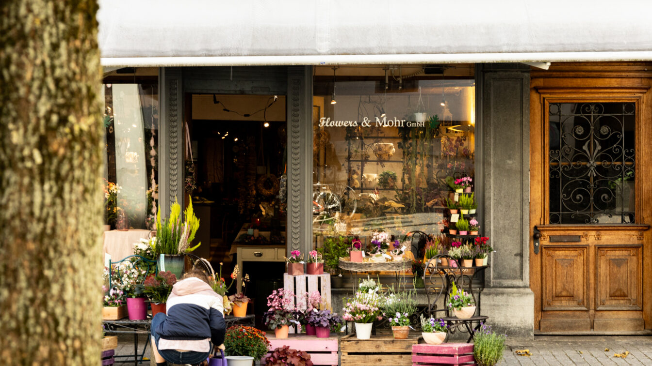 Ein gemütlicher Blumenladen in Berlin mit dem Namen ‚Flowers & Mohr GmbH‘. Vor dem Laden stehen bunte Blumentöpfe und Pflanzen auf Holzkisten und kleinen Tischen arrangiert. Eine Person kniet draußen und kümmert sich um die Pflanzen. Der Eingang ist von einer weißen Markise und einer dekorativen Holztür flankiert.