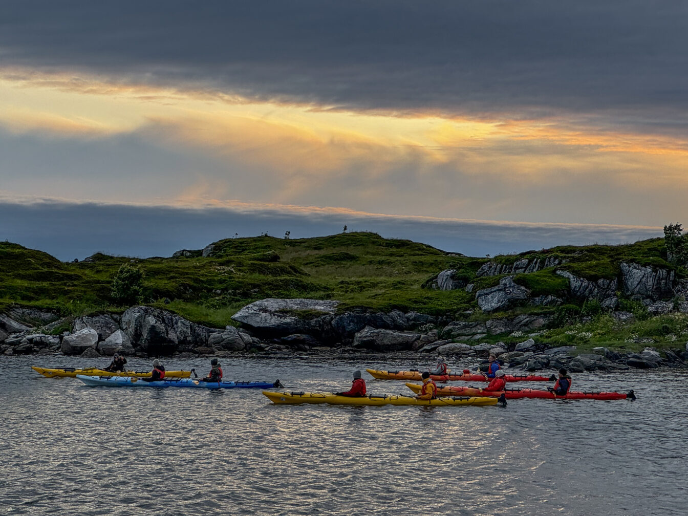 Gruppe von Reisenden paddelt gemeinsam in bunten Kajaks entlang der Küste der Lofoten bei Sonnenuntergang – aktive Gruppenreise in Norwegen mit Naturerlebnis.