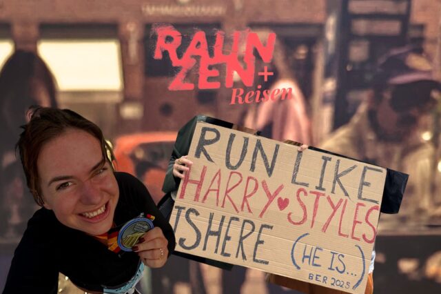Eine junge Frau lächelt glücklich in die Kamera und hält eine Medaille in der Hand. Daneben ist ein Pappschild zu sehen, auf dem steht: „RUN LIKE HARRY STYLES IS HERE (HE IS…) BER 2025“. Im Hintergrund verschwommen eine Straßenszene; darüber der Schriftzug „RAUNZEN + Reisen“ in rot