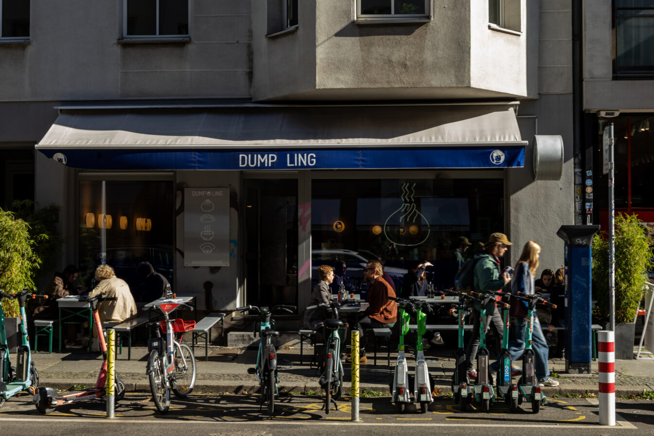 Straßenansicht eines Restaurants mit blauem Schild „DUMP LING“. Davor sitzen Menschen an Tischen und unterhalten sich. Mehrere E-Scooter und Fahrräder sind am Straßenrand geparkt. Die Szene wirkt lebendig und urban bei sonnigem Wetter.