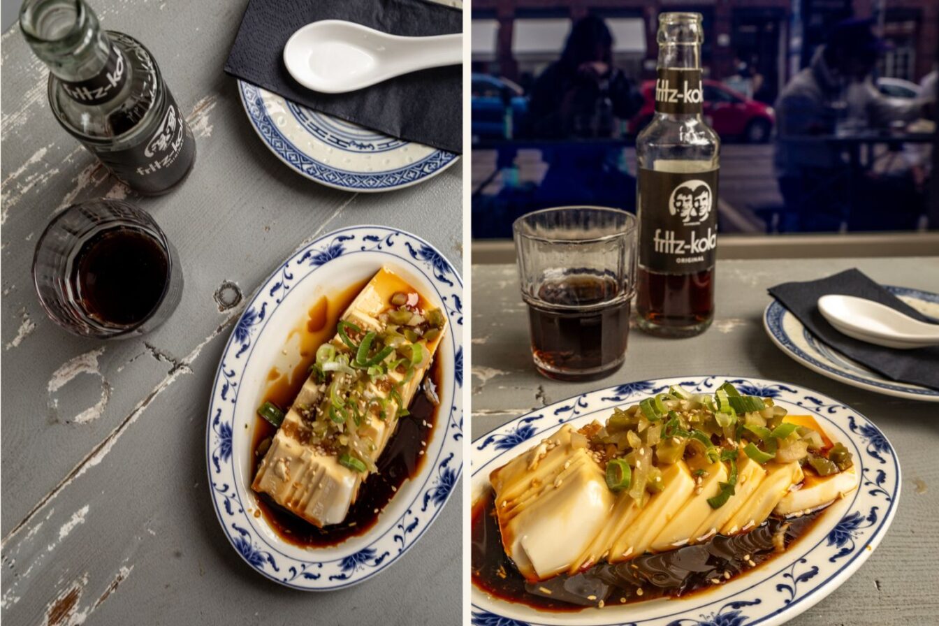 Zwei Ansichten desselben Gerichts: Ein rechteckiger Block Seidentofu auf einem Teller mit blauer Verzierung, serviert in Sojasauce, garniert mit Frühlingszwiebeln, Sesam und eingelegtem Gemüse. Daneben steht ein Glas mit dunkler Limonade und eine Flasche Fritz-Kola. Im Hintergrund sind Teller, Löffel und eine schwarze Serviette zu sehen.