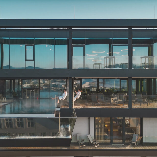 Zwei Personen in Bademänteln schaukeln auf der Dachterrasse eines Hotels, vor ihnen liegt ein Pool.