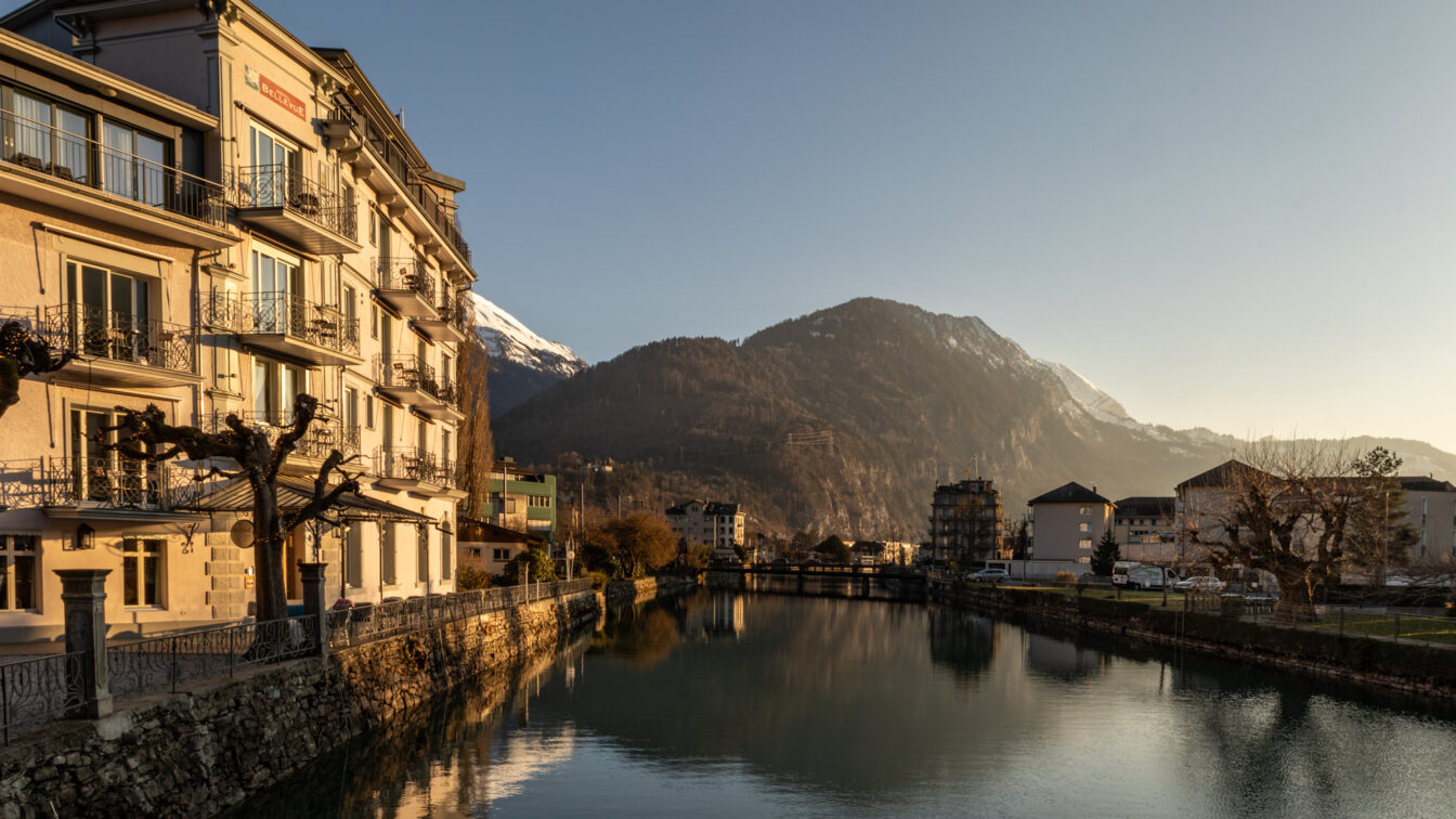 Herbstliche Abendstimmung in der Schweiz: Ein ruhiger Fluss spiegelt die umliegenden Häuser und schneebedeckten Berge, goldenes Sonnenlicht fällt auf die Fassaden und kahle Bäume am Ufer.