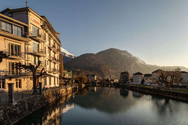 Herbstliche Abendstimmung in der Schweiz: Ein ruhiger Fluss spiegelt die umliegenden Häuser und schneebedeckten Berge, goldenes Sonnenlicht fällt auf die Fassaden und kahle Bäume am Ufer.